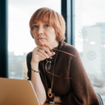 Loneliest-Job-in-the-Building-Manager-Portrait Thoughtful manager holding glasses while working at laptop by window